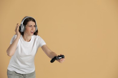 Woman in headphones playing video games with controller on beige background, space for text