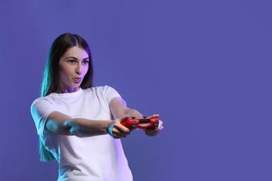 Surprised woman playing video games with controller on violet background, space for text