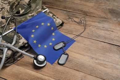 Stethoscope, flag of European Union, tags and military uniform on wooden table. Space for text