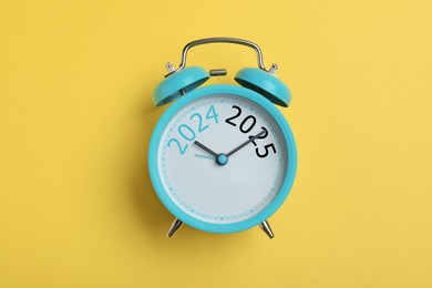 Alarm clock with hands pointing at numbers 2024 and 2025 on yellow background, top view. Beginning of new year