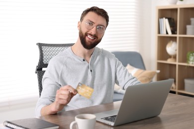 Photo of Online banking. Happy young man with credit card and laptop paying purchase at table indoors