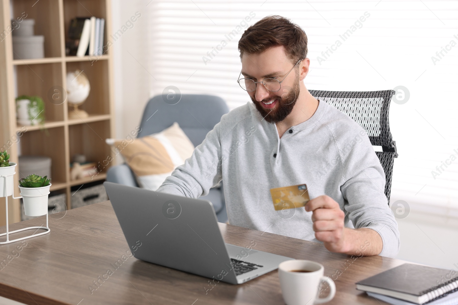 Photo of Online banking. Happy young man with credit card and laptop paying purchase at table indoors