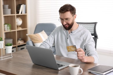 Online banking. Happy young man with credit card and laptop paying purchase at table indoors