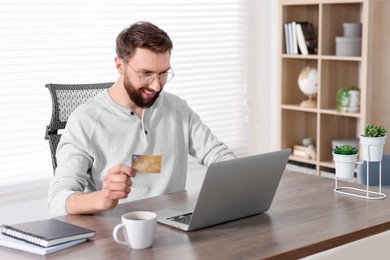 Online banking. Happy young man with credit card and laptop paying purchase at table indoors