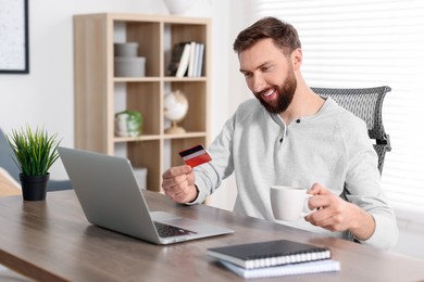 Photo of Online banking. Happy young man with credit card and laptop paying purchase at table indoors