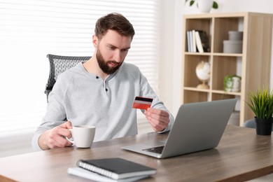 Photo of Online banking. Young man with credit card and laptop paying purchase at table indoors