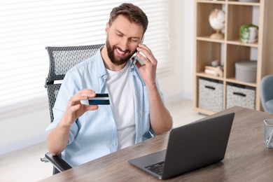 Photo of Online banking. Happy young man with credit card talking by smartphone at table indoors