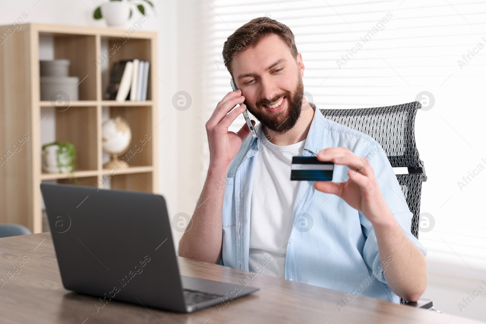 Photo of Online banking. Happy young man with credit card talking by smartphone at table indoors