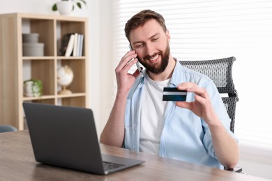 Photo of Online banking. Happy young man with credit card talking by smartphone at table indoors