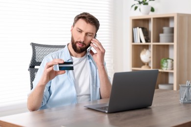 Online banking. Young man with credit card talking by smartphone at table indoors