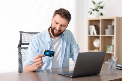 Photo of Online banking. Happy young man with credit card and laptop paying purchase at table indoors