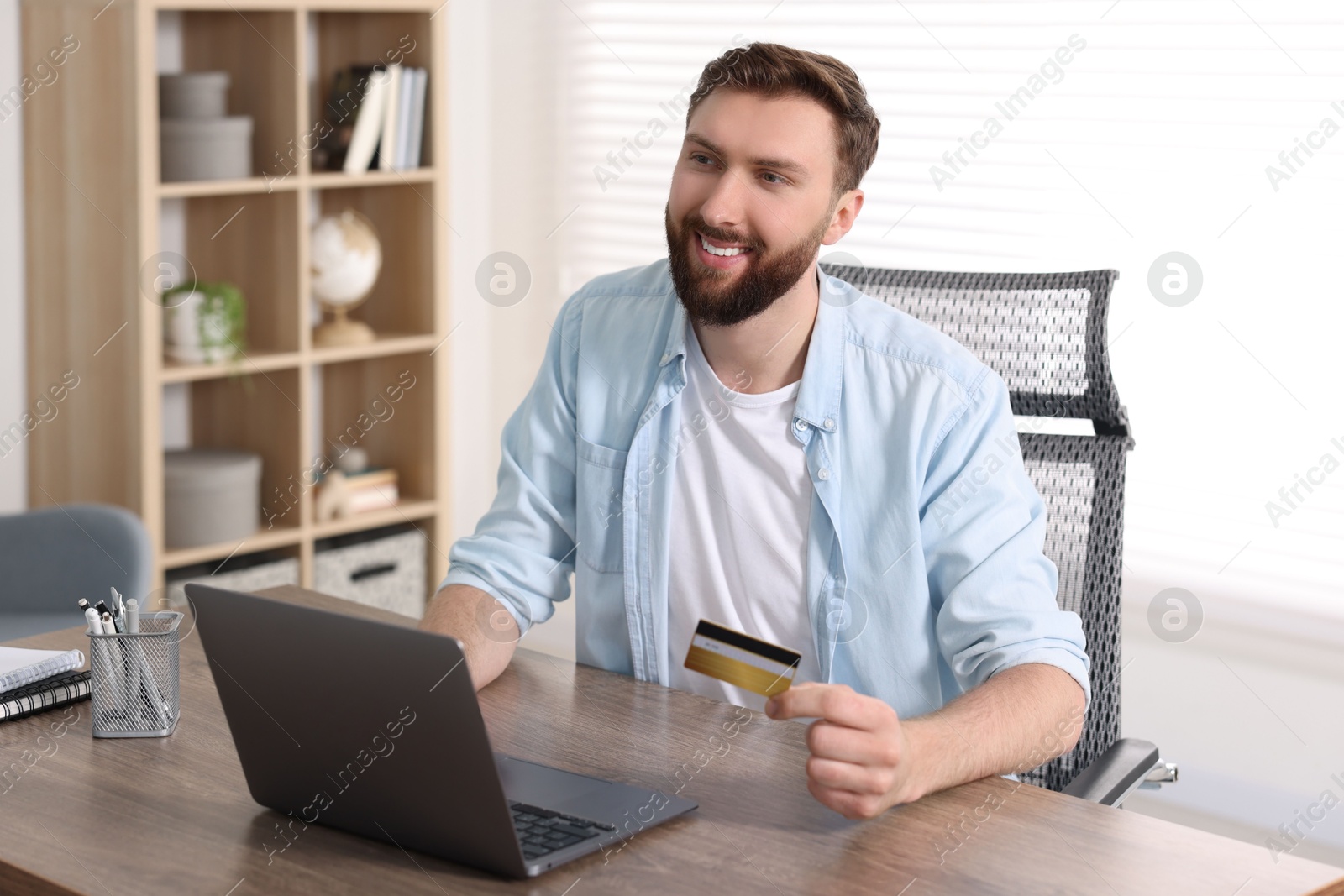 Photo of Online banking. Happy young man with credit card and laptop paying purchase at table indoors