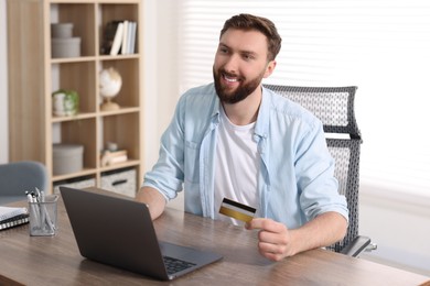 Photo of Online banking. Happy young man with credit card and laptop paying purchase at table indoors