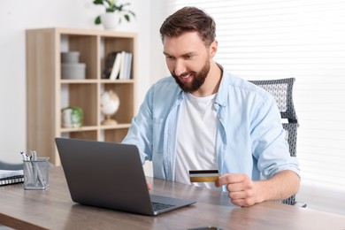 Photo of Online banking. Happy young man with credit card and laptop paying purchase at table indoors