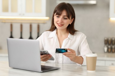 Photo of Online banking. Smiling woman with credit card, smartphone and laptop paying purchase at table indoors