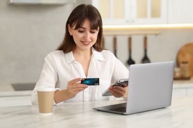 Online banking. Smiling woman with credit card, smartphone and laptop paying purchase at table indoors