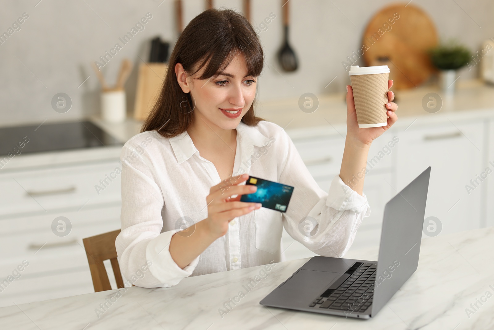 Photo of Online banking. Smiling woman with credit card, paper cup and laptop paying purchase at table indoors