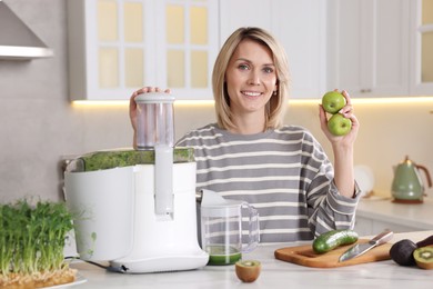 Smiling woman with juicer and fresh products at white marble table in kitchen