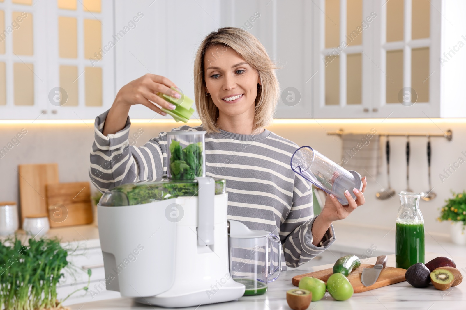 Photo of Smiling woman putting fresh celery into juicer at white marble table in kitchen