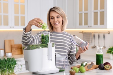 Smiling woman putting fresh celery into juicer at white marble table in kitchen