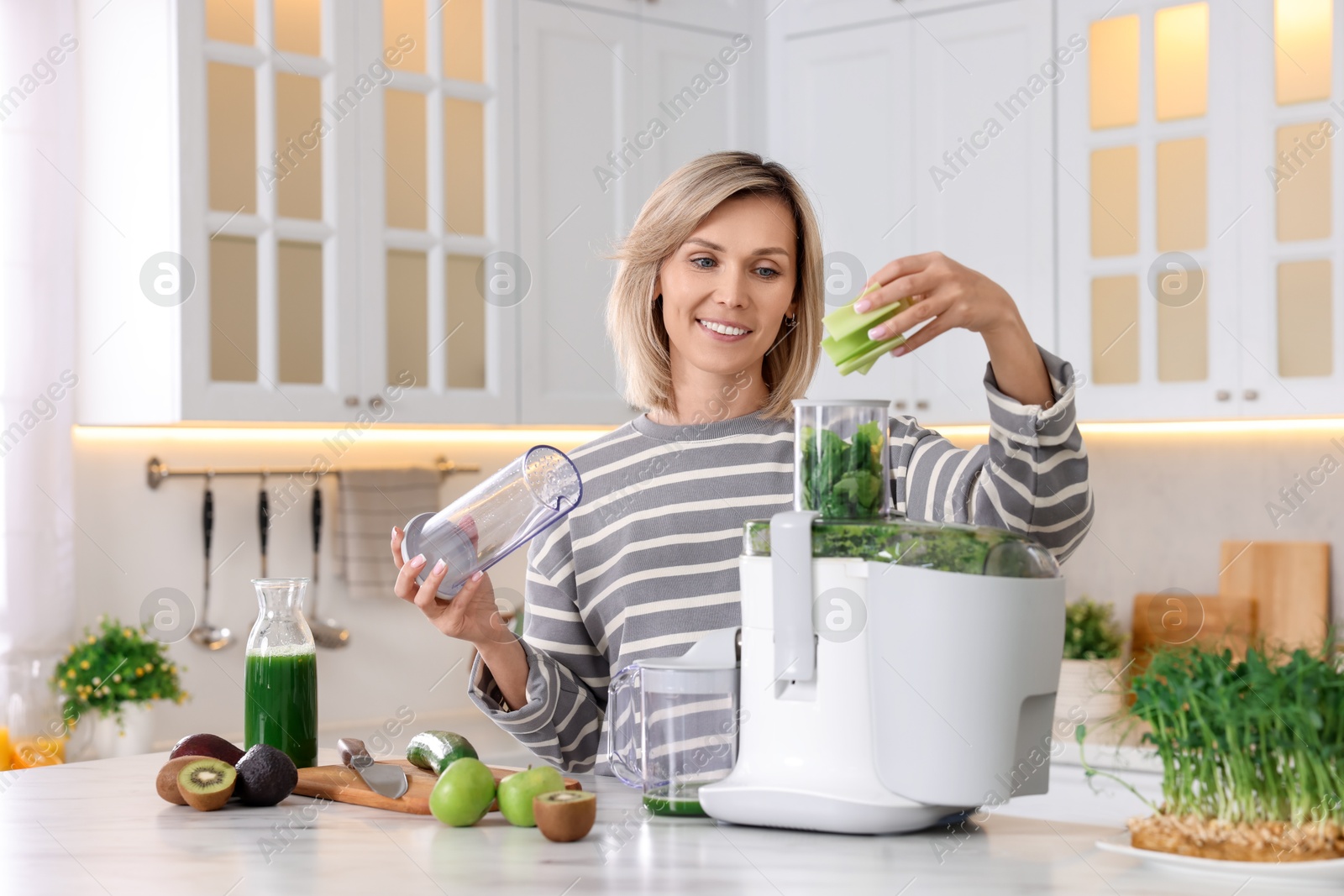 Photo of Smiling woman putting fresh celery into juicer at white marble table in kitchen