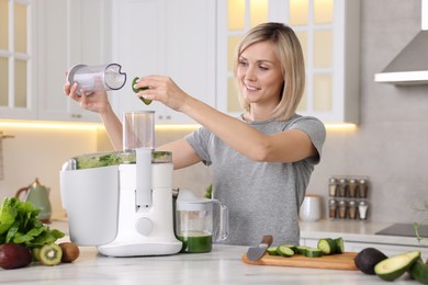 Smiling woman putting fresh cucumber into juicer at white marble table in kitchen