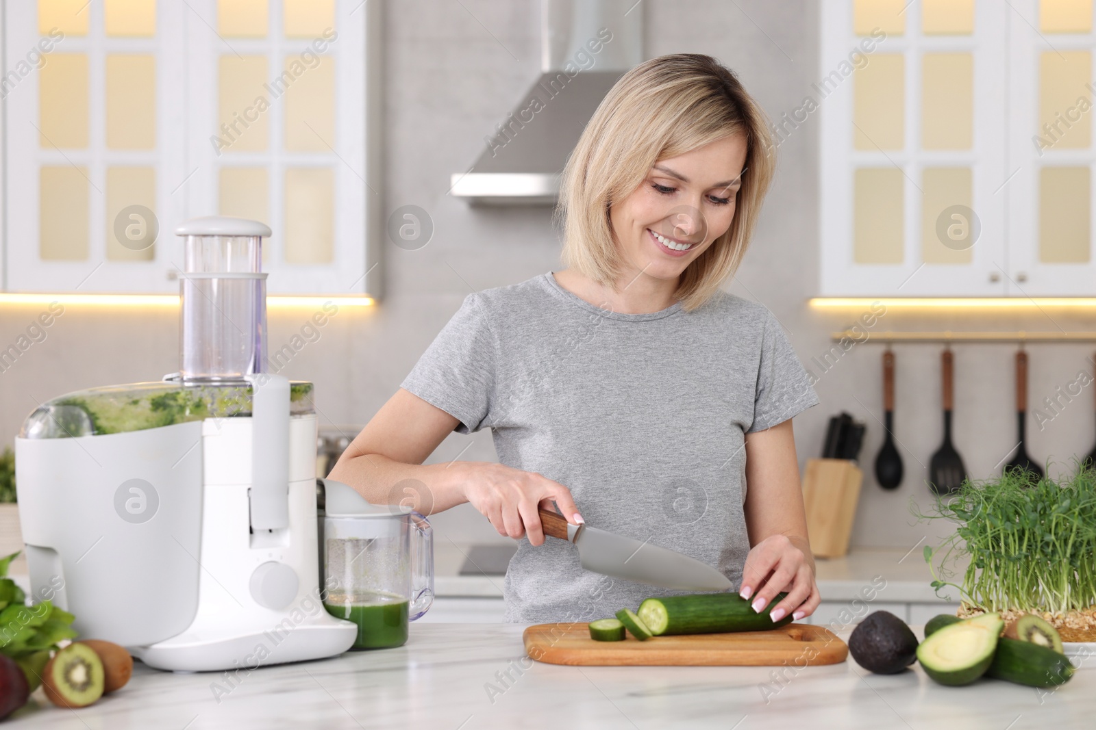 Photo of Juicer and fresh products on white marble table. Smiling woman cutting cucumber in kitchen
