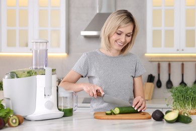 Juicer and fresh products on white marble table. Smiling woman cutting cucumber in kitchen