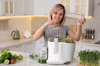 Smiling woman putting fresh basil into juicer at white marble table in kitchen