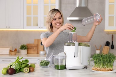 Photo of Smiling woman putting fresh basil into juicer at white marble table in kitchen