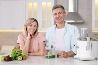 Happy couple with juicer, fresh products and tasty juice at white marble table in kitchen