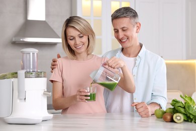 Happy couple with juicer, fresh products and tasty juice at white marble table in kitchen