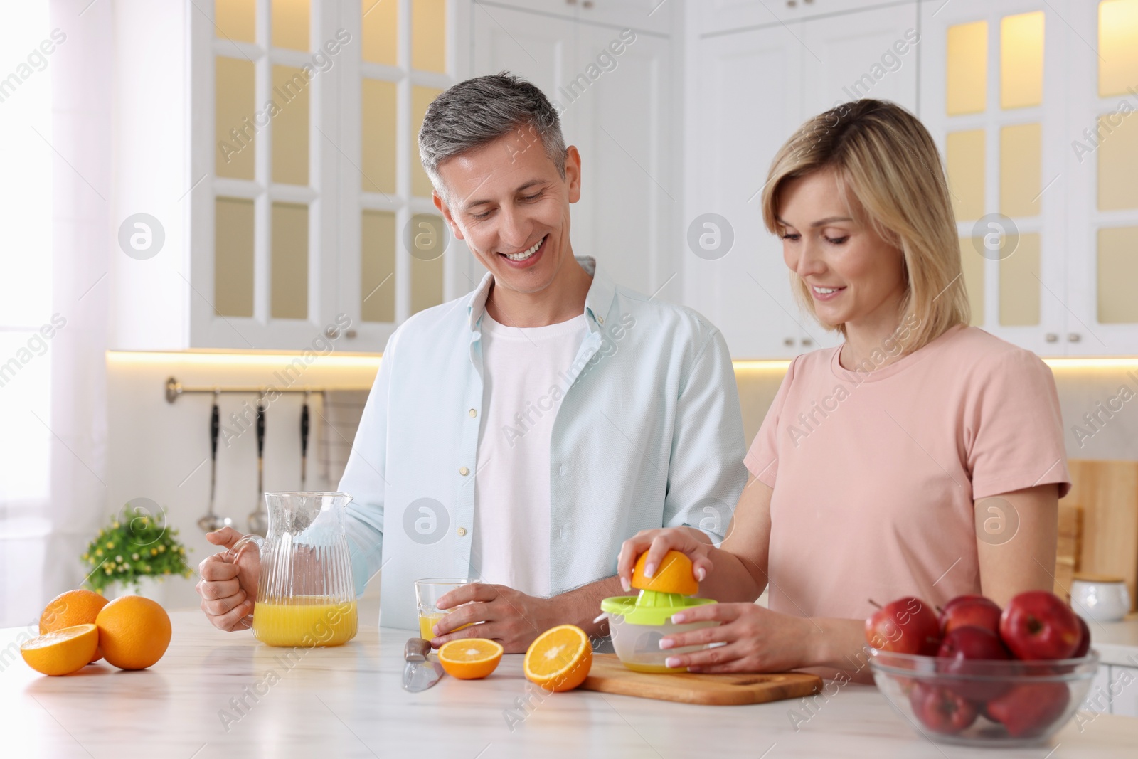 Photo of Happy couple with juicer and fresh products making juice at white marble table in kitchen