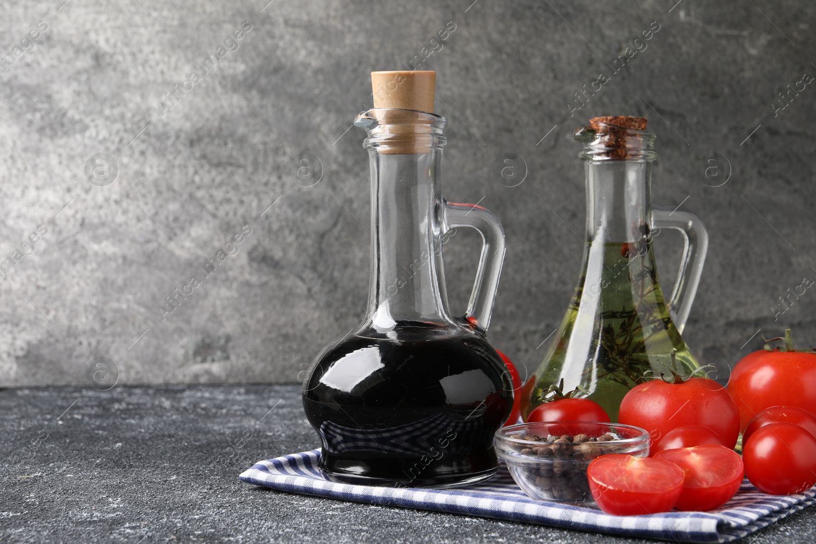 Photo of Balsamic vinegar in glass jug, oil, tomatoes and spices on gray textured table, space for text