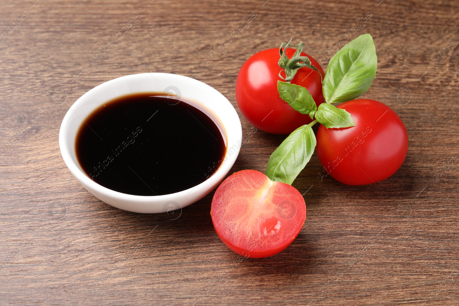 Photo of Balsamic vinegar in bowl, basil and tomatoes on wooden table