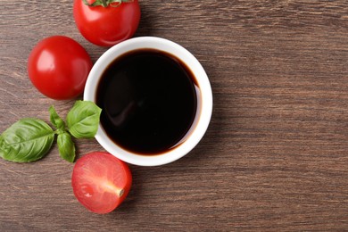 Balsamic vinegar in bowl, basil and tomatoes on wooden table, flat lay. Space for text