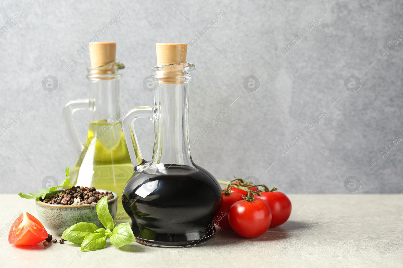 Photo of Balsamic vinegar in glass jug, oil, tomatoes, herbs and spices on gray textured table, space for text