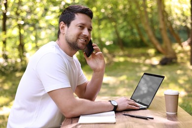 Photo of Smiling freelancer talking on smartphone at table with laptop outdoors. Remote job