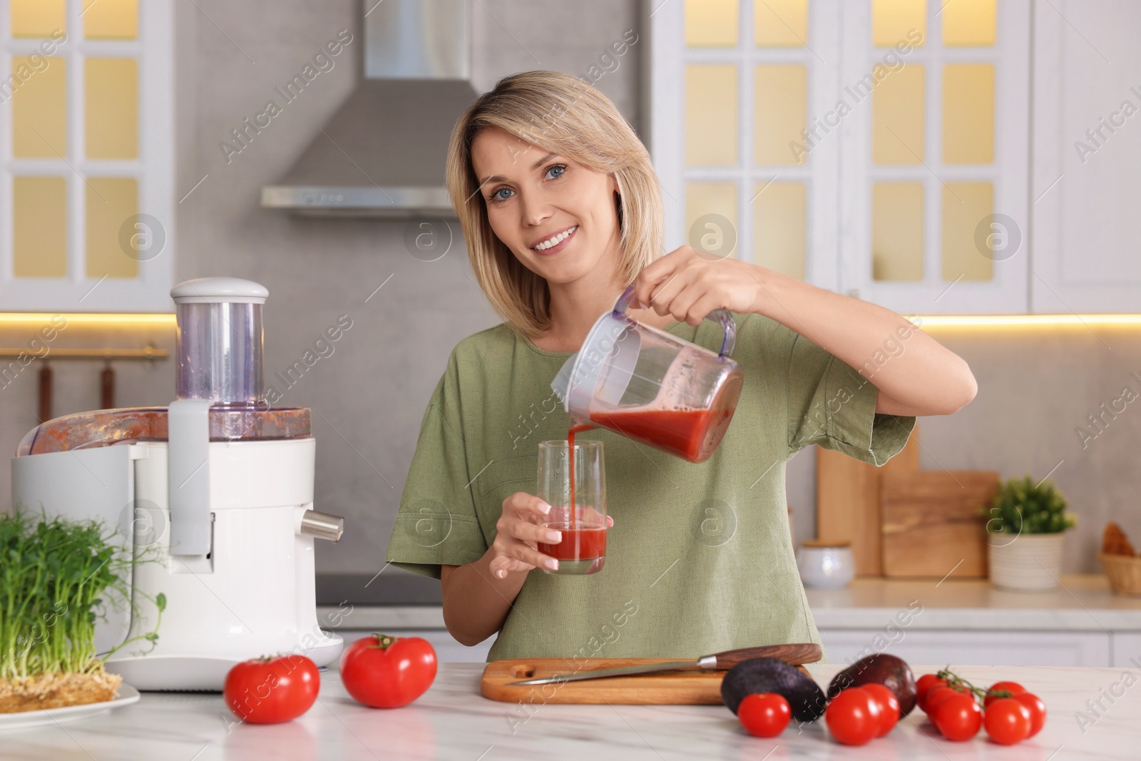 Photo of Smiling woman pouring tomato juice into glass in kitchen. Juicer and fresh products on white marble table