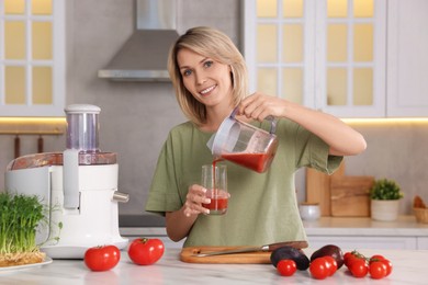 Smiling woman pouring tomato juice into glass in kitchen. Juicer and fresh products on white marble table