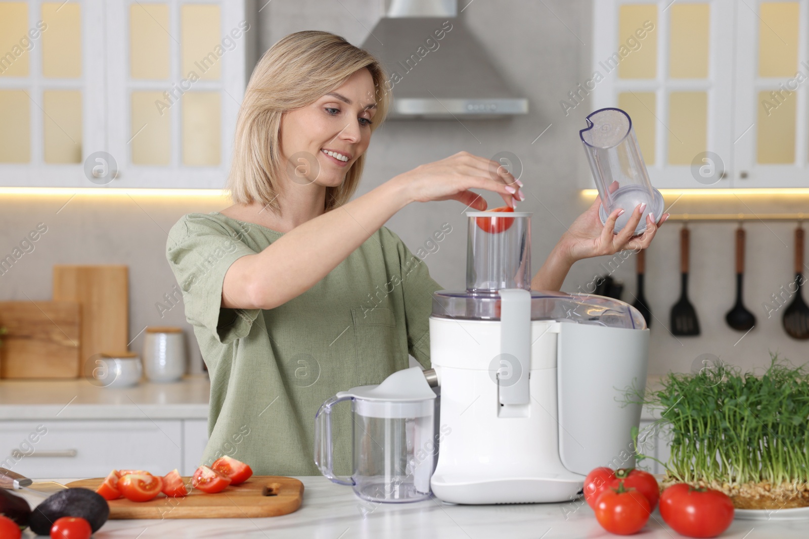 Photo of Smiling woman putting fresh tomato into juicer at white marble table in kitchen