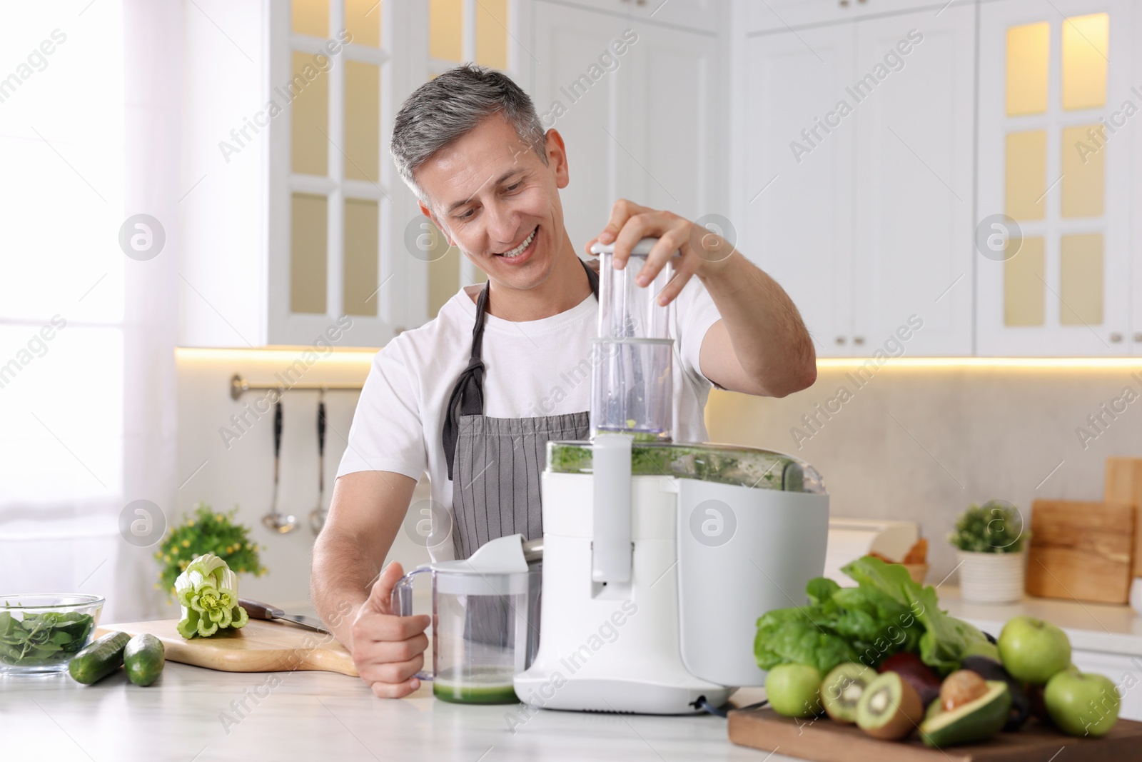 Photo of Smiling man with fresh products using juicer at white marble table in kitchen