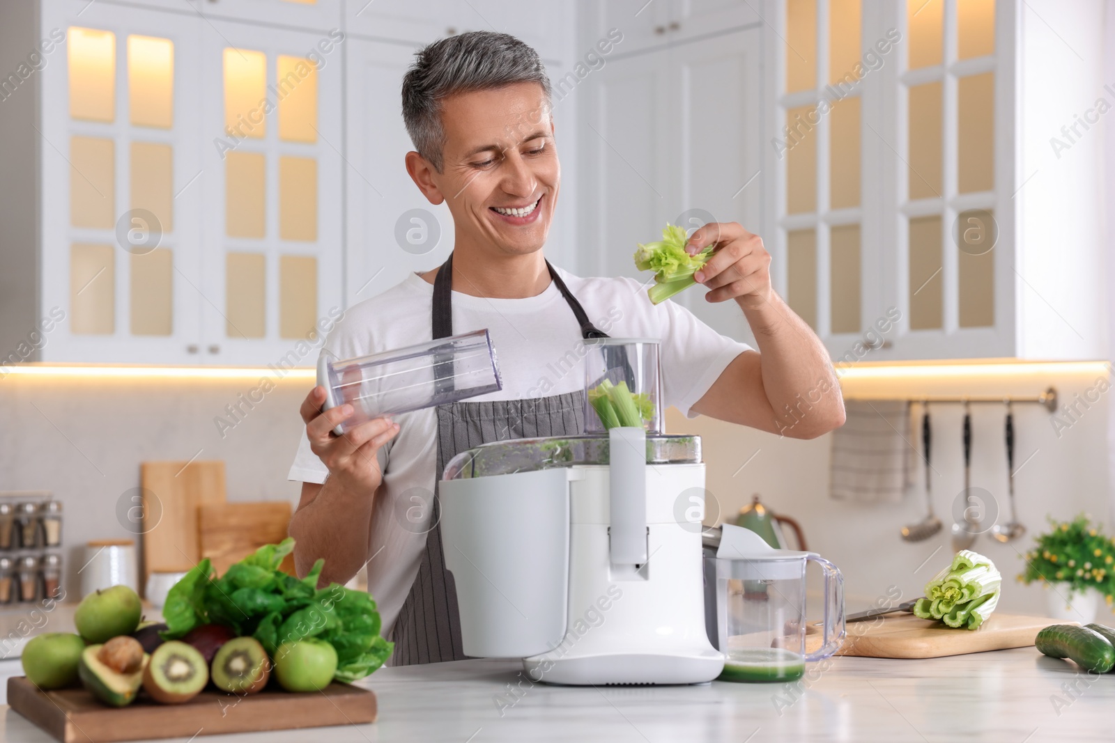 Photo of Smiling man putting fresh celery into juicer at white marble table in kitchen