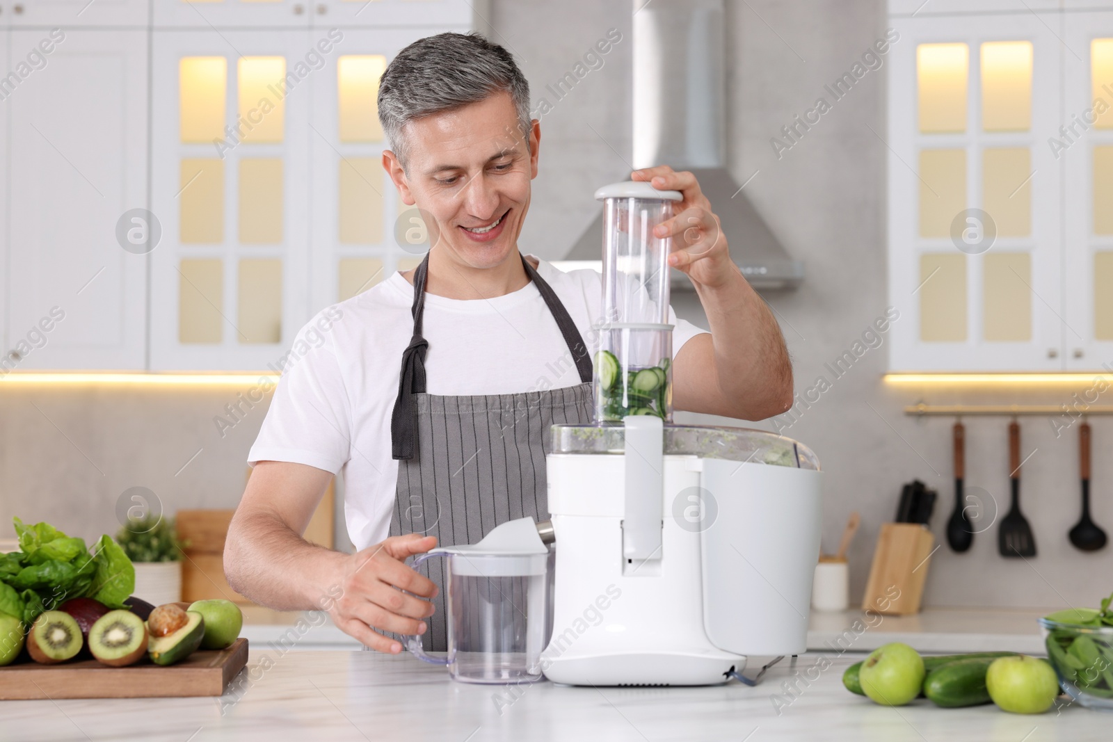 Photo of Smiling man with fresh products using juicer at white marble table in kitchen