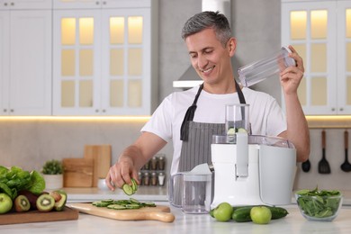 Smiling man putting fresh cucumber into juicer at white marble table in kitchen