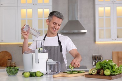 Photo of Smiling man putting fresh cucumber into juicer at white marble table in kitchen