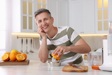 Smiling man squeezing fresh orange with juicer at white marble table in kitchen