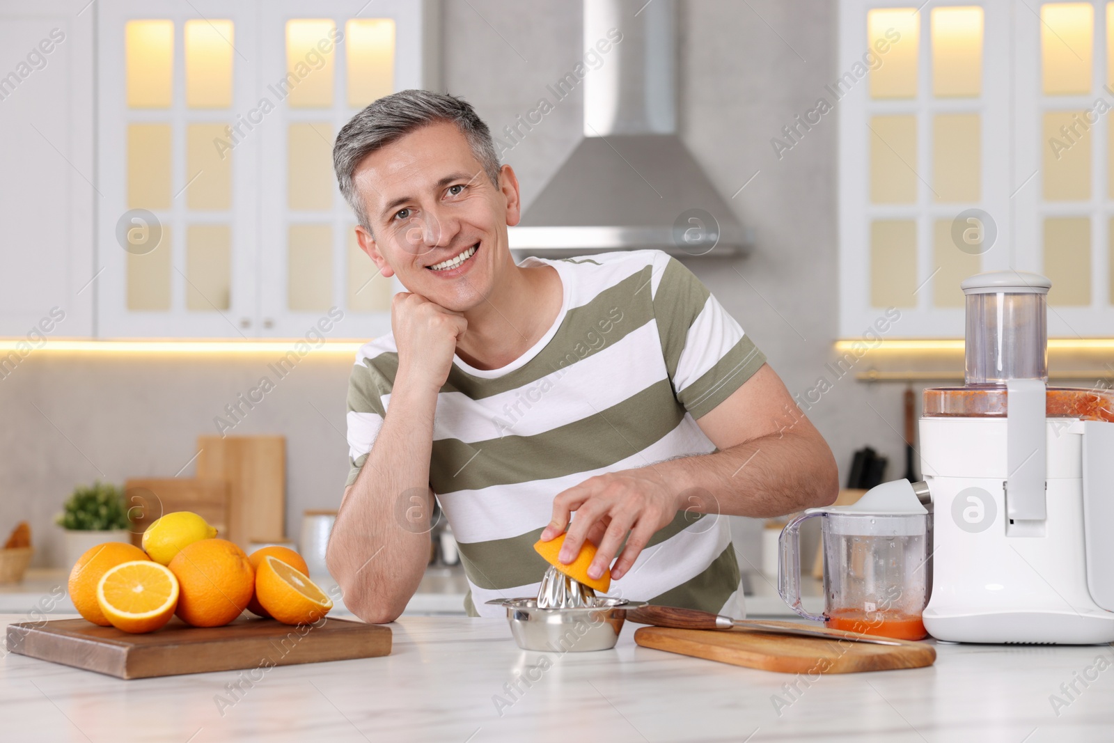 Photo of Smiling man squeezing fresh orange with juicer at white marble table in kitchen