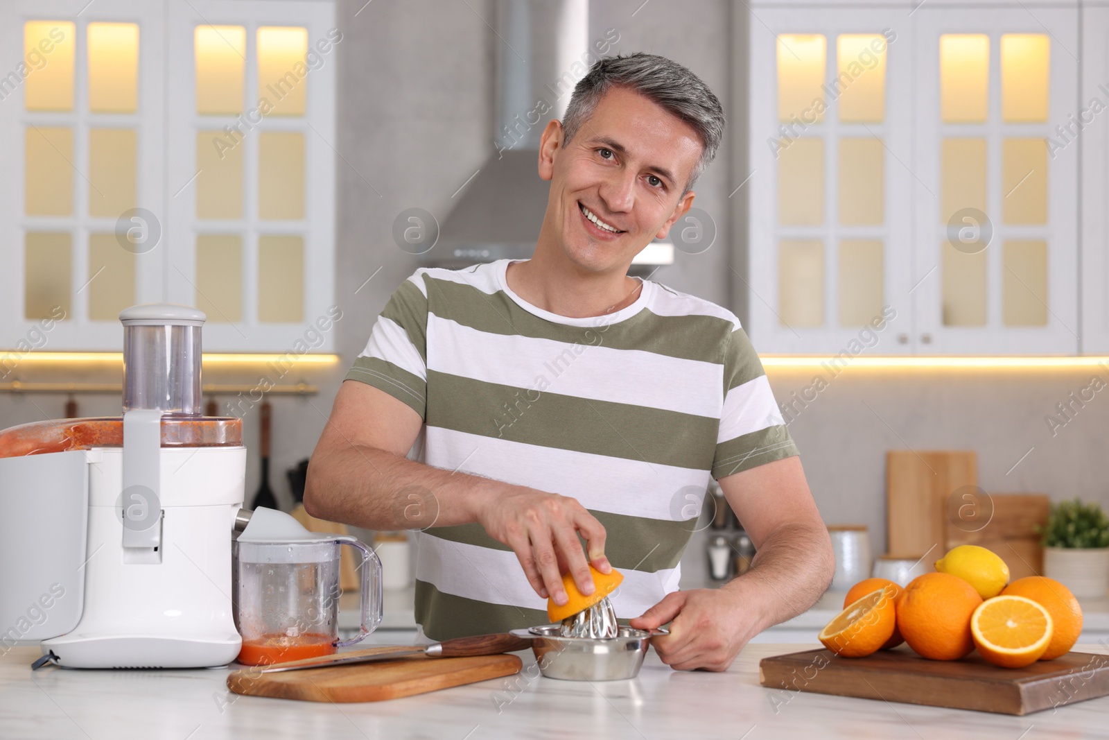 Photo of Smiling man squeezing fresh orange with juicer at white marble table in kitchen