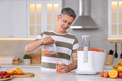 Photo of Smiling man pouring juice into glass in kitchen. Juicer and fresh products on white marble table